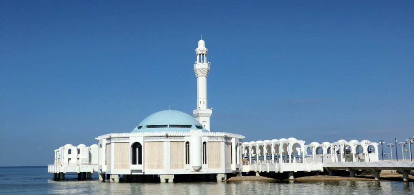 Mosquée blanche sur pilotis, dôme bleu, ciel clair.